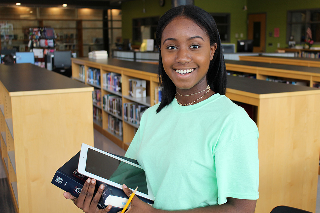 Jasmine standing in a library holding a book and tablet