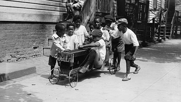 a group of African-American children playing with a pedal car along a city street