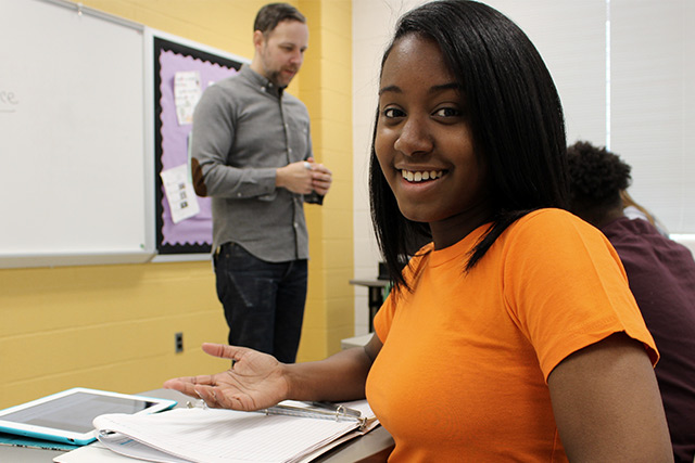 Jasmine talking to the camera with Mr. Lexington in the background talking to class